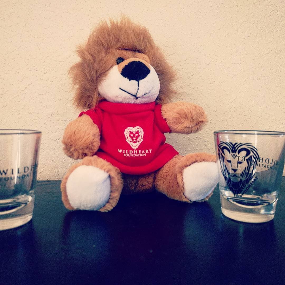 A small lion stuffy wearing a t-shirt with the WildHeart logo, next to two shot glasses that also have the WildHeart logo on it